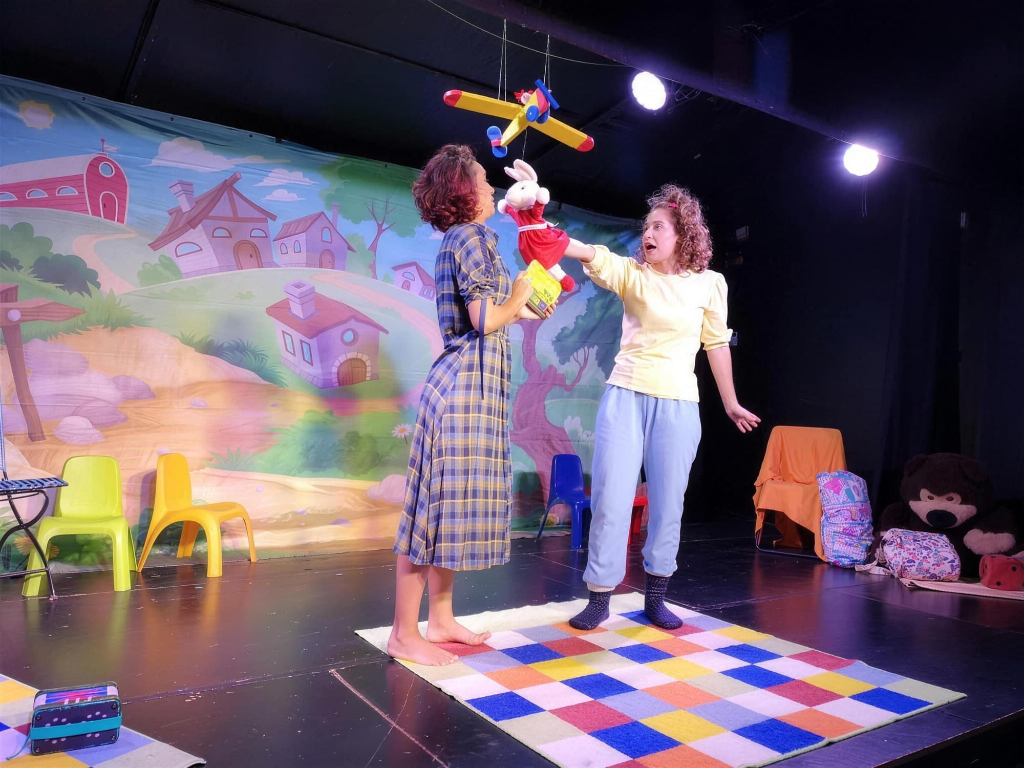 שתי שחקניות על במה צבעונית עם בובת ארנב ומטוס צעצוע תלוי, בהצגה לקטנטנים בתיאטרון הפארק